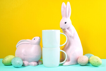 Easter two white coffee mugs with pink bunnies and easter eggs on yellow and blue background. Negative copy space for your text or design here.