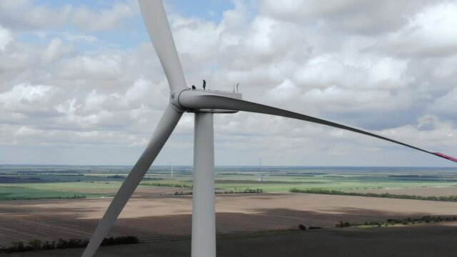 Skilled workers walk on top service ground of windmill with blades against sky with fluffy white clouds on sunny day aerial view