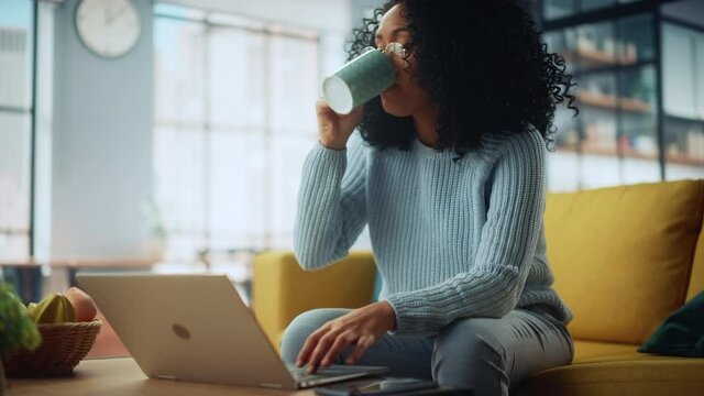 Beautiful Authentic Latina Female Sitting on a Sofa in a Cozy Living Room and Using Laptop Computer at Home. She's Drinking from a Cup and Browsing Internet and Social Networks.