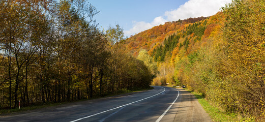 Zakarpattia Oblast, Ukraine - October 27, 2020: Among the mountains, on the side of a deserted asphalt road, there is a parked car. Autumn trees all around, yellowed and reddened leaves. Blue sky.