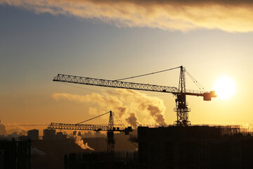 Silhouettes of construction cranes and unfinished residential buildings on sunrise sky and smoke background. Housing construction, apartment block in city