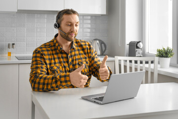 A man during an online video call. He sits at home in front of a laptop monitor and showing thumbs up gesture.