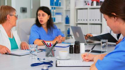 Nurse writing on clipboard while profesional teamworkers having medical meeting discussing in background in brainstorming office. Profesional doctors examining the symptoms of patient in meeting room.