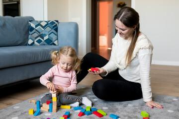 Mother and daughter playing wooden cubes on the floor of the living room at home. Activities with children inside, early childhood development