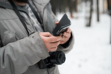 Photographer uses mobile phone outside in winter. Man in a jacket and with a camera, close-up. Selective focus