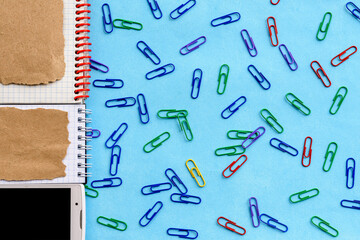 Top view of an office desk with an empty notepad, smartphone, and paper clips on a blue background