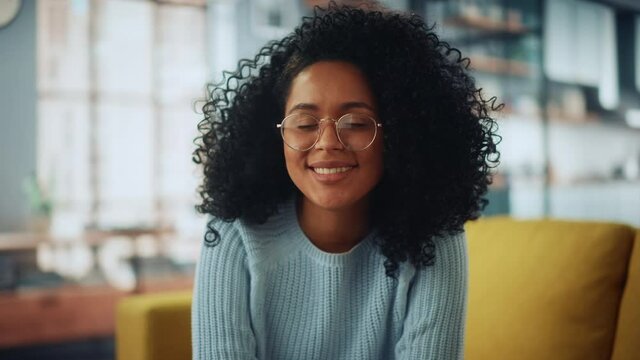 Portrait of a Beautiful Authentic Latina Female with Afro Hair Wearing Light Blue Jumper and Glasses. She Looks to the Camera and Smiling Charmingly. Successful Woman Resting in Bright Living Room.