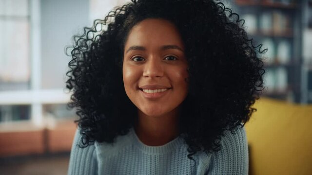 Portrait of a Beautiful Authentic Latina Female with Stylish Afro Hair Wearing Light Blue Jumper. She Looks to the Camera and Smiling Charmingly. Successful Woman Resting in Bright Living Room.