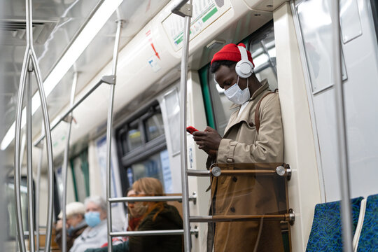 Afro-American hipster man standing in subway train, wear face medical mask to protect yourself from contact with flu virus, covid-19, using cellphone, listens to music with wireless headphones. 