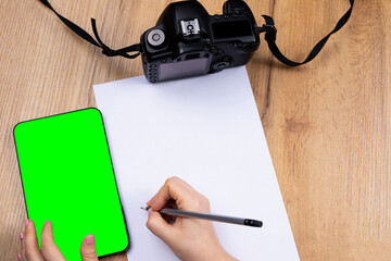 Flat lay, hands writing in a blank white page from digital mock up green screen tablet, decorated with vintage camera for photography tool on table wooden background