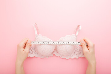 Young adult woman hands holding white measure tape and measuring bra on light pink table background. Pastel color. Closeup. Point of view shot. Choosing right size. Top down view.