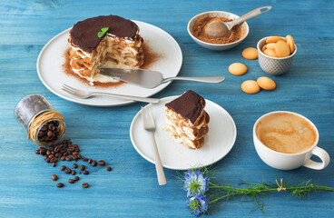 Round tiramisu, homemade no-bake traditional Italian sweet dessert. Blue table with cake, bowl with cocoa powder, round sponge biscuits, coffee beans and coffee with milk. Blue background.