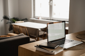 Fototapeta premium Laptop computer with blank screen, notebooks on wooden table in warm sunlight room. Aesthetic minimal interior design template with mockup copy space. Blog, web, social media concept.