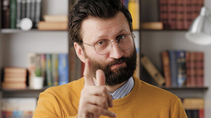 Man threatens with his finger. Serious frowning bearded man in glasses in office or apartment room looking at camera and points menacingly with his index finger. Close up