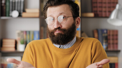 Shrug, i dont know. Perplexed bearded man in glasses in office or apartment room looking at camera...