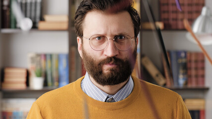 Fototapeta premium Angry man, rage. Angry annoyed bearded man in glasses in office or apartment room looking at camera and hits table with his fist, pencils fly to sides. Close-up