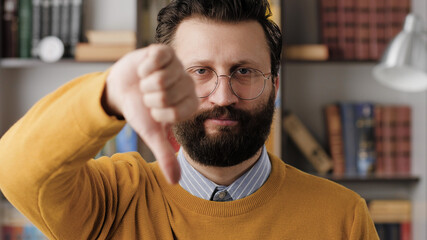 Man thumbs down. Serious displeased bearded man with glasses in office or apartment room looking at camera and shows his hand with thumb down. Close-up