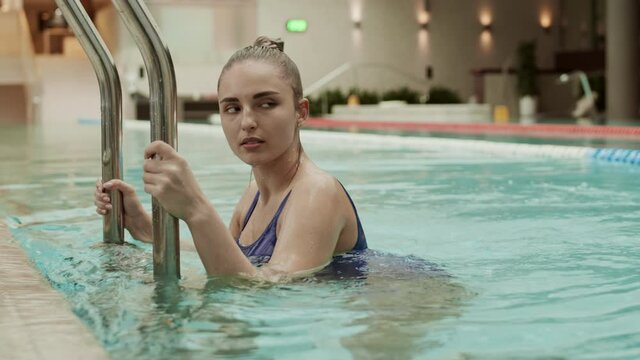 Medium shot portrait of young Caucasian woman with blond hair wearing blue swimming suit swimming in pool in luxury hotel and looking at camera