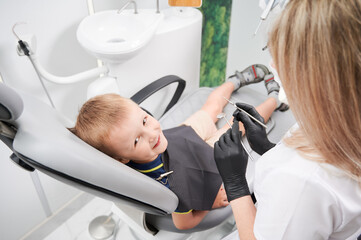 Adorable male child looking at camera and smiling while female dentist holding dental mirror and explorer in dental office. Concept of pediatric dentistry and dental care.