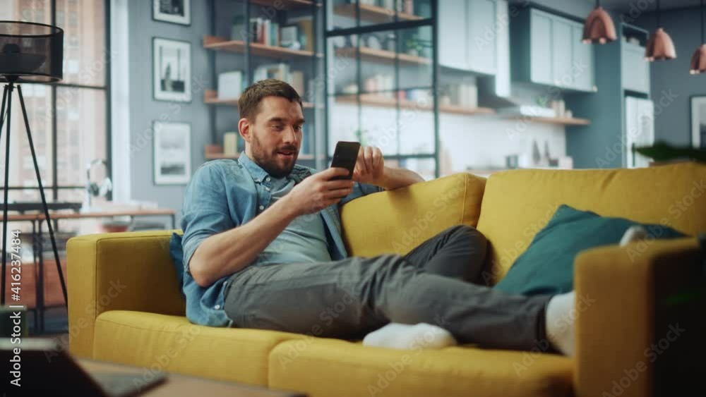 Leinwandbilder happy handsome caucasian man using smartphone in cozy living room at home. man resting on comfortabl