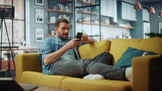Happy Handsome Caucasian Man Using Smartphone in Cozy Living Room at Home. Man Resting on Comfortable Sofa. He's Browsing the Internet and Checking Videos on Social Networks and Having Fun.