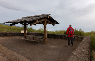 An elderly hiker with a backpack stands in the early morning at a lookout point on the mountain on the north side of the Atlantic island of Tenerife. He looks down at the clouds and the Atlantic Ocean