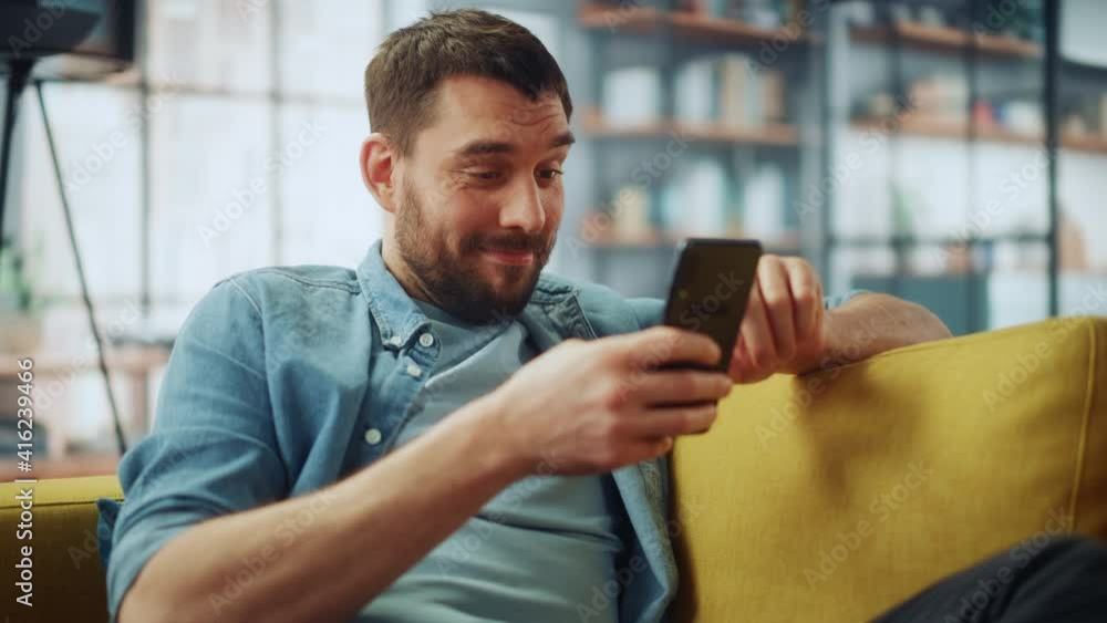 Aufkleber Happy Handsome Caucasian Man Using Smartphone in Cozy Living Room at Home. Man Resting on Comfortable Sofa. He's Browsing the Internet and Checking Videos on Social Networks and Having Fun.