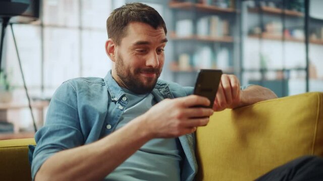 Happy Handsome Caucasian Man Using Smartphone in Cozy Living Room at Home. Man Resting on Comfortable Sofa. He's Browsing the Internet and Checking Videos on Social Networks and Having Fun.