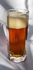 Semi-dark beer and foam in a faceted glass glass close-up, illuminated by the sun on a white background