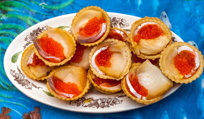 Obraz na płótnie Canvas Tartlets with oily fish and red caviar on a ceramic plate close up on the background of a blue tablecloth with a picture of fish