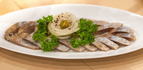 Sliced salted fish herring decorated with a sprig of parsley, onion rings and green olive on an oval white ceramic plate close-up on a tablecloth background