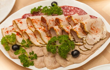 Sliced boiled meat, sausage, black olives and sprigs of parsley close-up on a white plate on a tablecloth background