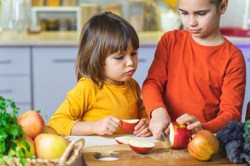 Fototapeta premium Funny kids cutting apples in kitchen, having fun. Happy sister and brother cook together laughing, healthy juices and fresh fruit salad. Fun vegan family cooking events