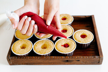 the hands of a female pastry chef hold a cooking bag and fill the red berry filling cupcakes in a wooden tray.Food for breakfast. Freshly baked cupcakes for dessert. Foodies and cuisine.