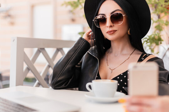 Fashionable woman in elegant hat in fashion sunglasses in trendy clothes sits and makes selfie on smartphone in cafe. Stylish girl in black youth wear photographs herself on summer poen terrace.