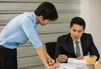 Asian senior good-looking boss in suit sitting at desk and working with junior employee in office with roll of paper. Businessmen teamwork and good colleagues concept