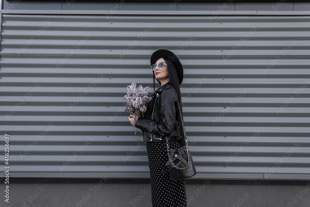 Wall mural Fashionable model woman in stylish sunglasses in hat in black youth outfit stands with bouquet of lilac flowers near metal vintage wall in city. Attractive girl enjoys walk and fresh amazing flowers.