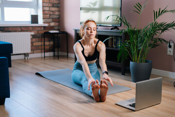 Naklejka na ściany i meble Sporty redhead young woman working out, doing stretching exercise on yoga mat while watching fitness video online on laptop at home. Concept of sports training red-haired female during quarantine.