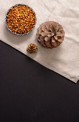 A bowl of peeled pine nut kernels and two pine cones on a linen cloth and a black table.