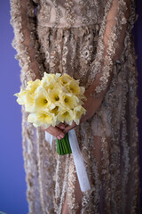 Closeup portrait of a bride in beautiful cream wedding dress, standing on a violet background and holding a bouquet of yellow flowers, and greenery with a white ribbon