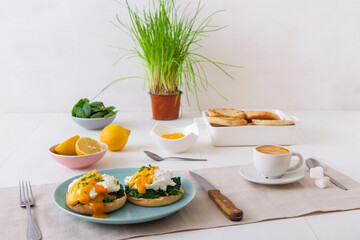 Eggs Florentine, English muffins, spinach, poached eggs, Mornay sauce, chive herbs, lemon, cup of coffee. White wooden table with breakfast.