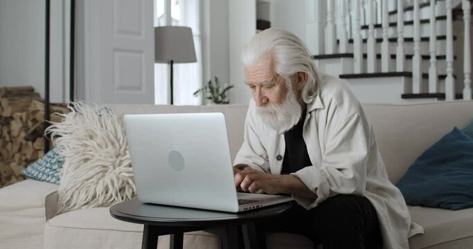 Crop view of elderly grey haired grandfather typing while communicating online. Male bearded retiree using laptop while sitting on sofa at home. Concept of leisure and retirement