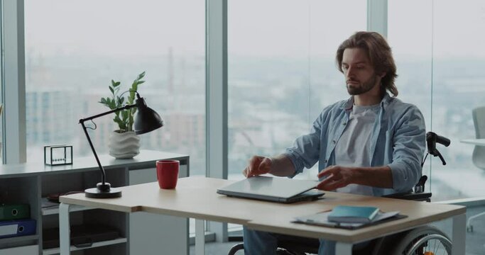 Young caucasian man with disabilities sitting in wheelchair using laptop computer inside corporate office shared workspace indoors.