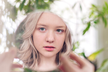 The reflection in the mirror of a beautiful blonde girl looking at herself. Plant leaves in the background. Psychological concept. Urban Jungle concept.