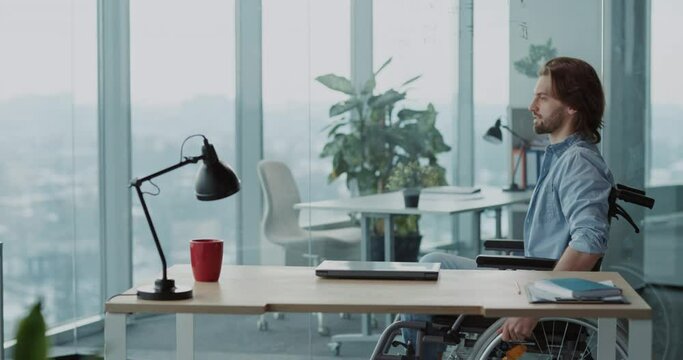 Corporate young man disabled in black wheelchair moving to desk with laptop opening device connecting network starting work in modern company.
