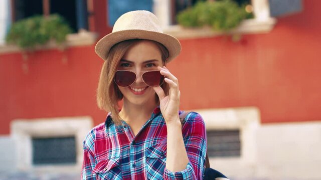 Portrait of young beautiful girl tourist in sunglasses and hat standing on background of bright houses and smiling posing at camera.