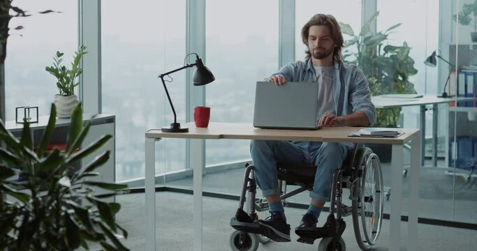 Young corportae confident man person with disabilities sitting in wheelchair performing online working project via laptop in modern company.