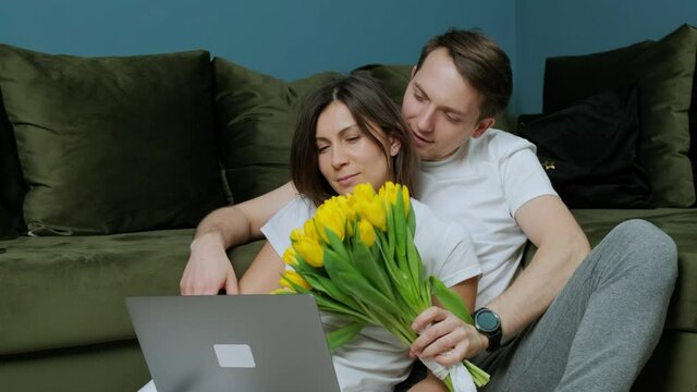 Young woman that is working at the computer receiving a bouquet of yellow tulips from her husband and kissing: romance, anniversaries.
