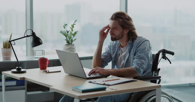 Young office executive handsome disabled male worker in wheelchair sitting by computer desk doing work inside business company.