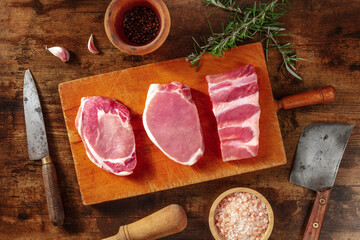 Pork meat, various cuts, shot from the top on a dark rustic wooden background with rosemary, salt, and pepper